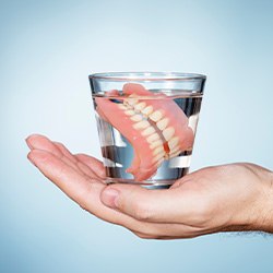 Full set of dentures in a glass of clear fluid resting in the palm of a hand with a grey-blue background